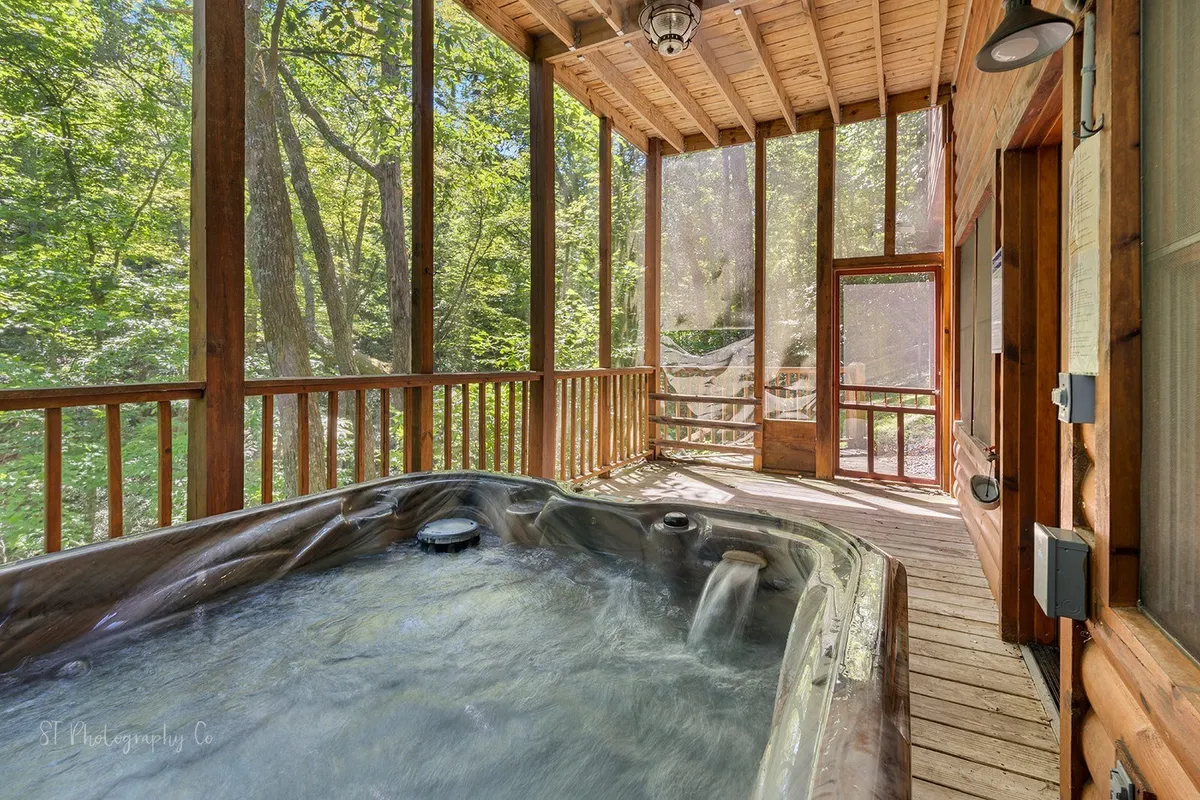 An inviting indoor hot tub set against a backdrop of large windows and a wooden deck, seamlessly blending the comfort of the interior with the beauty of the natural surroundings. The space encourages relaxation and social gatherings, with views of the trees and sunset filtering through the windows, creating a calming and rejuvenating atmosphere.