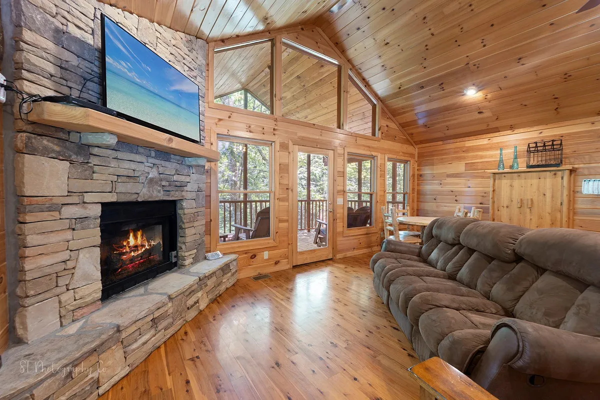 A cozy, inviting living area with a gas fireplace, large windows revealing wooded views, and comfortable seating. The warm ambiance is perfect for couples to snuggle up, enjoy a warm drink, or watch the sunset together.