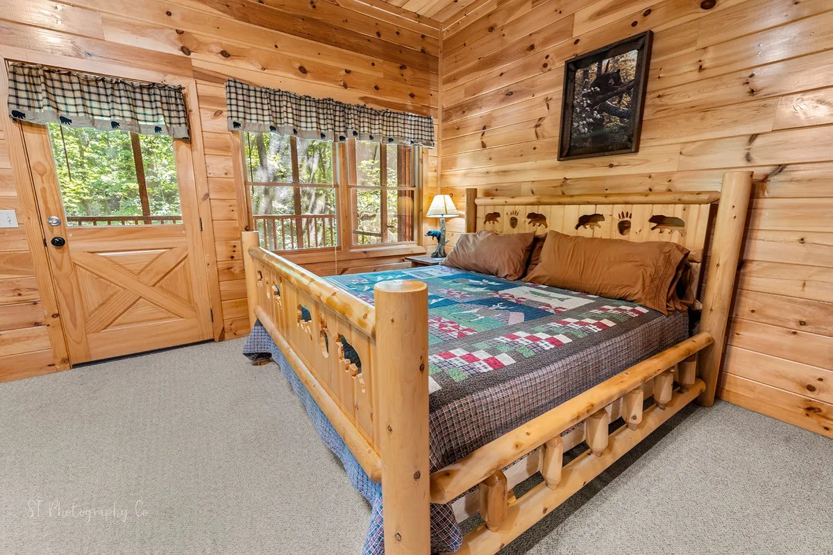 A peaceful bedroom with a log bed adorned with a colorful quilt, surrounded by large windows and a wooden door that frame serene views of nature. The setting is tranquil, perfect for soaking in the sights of changing leaves or a quiet evening after a day spent outdoors.