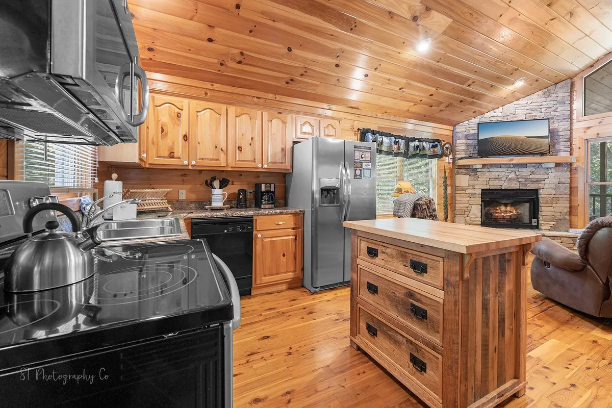 A warm and welcoming kitchen scene featuring a spacious kitchen island, stainless steel refrigerator, and wooden cabinets. The open layout is perfect for cooking, entertaining, and gathering with family. The cozy fireplace adds to the inviting atmosphere, while the space is clearly designed for preparing meals and spending quality time together.