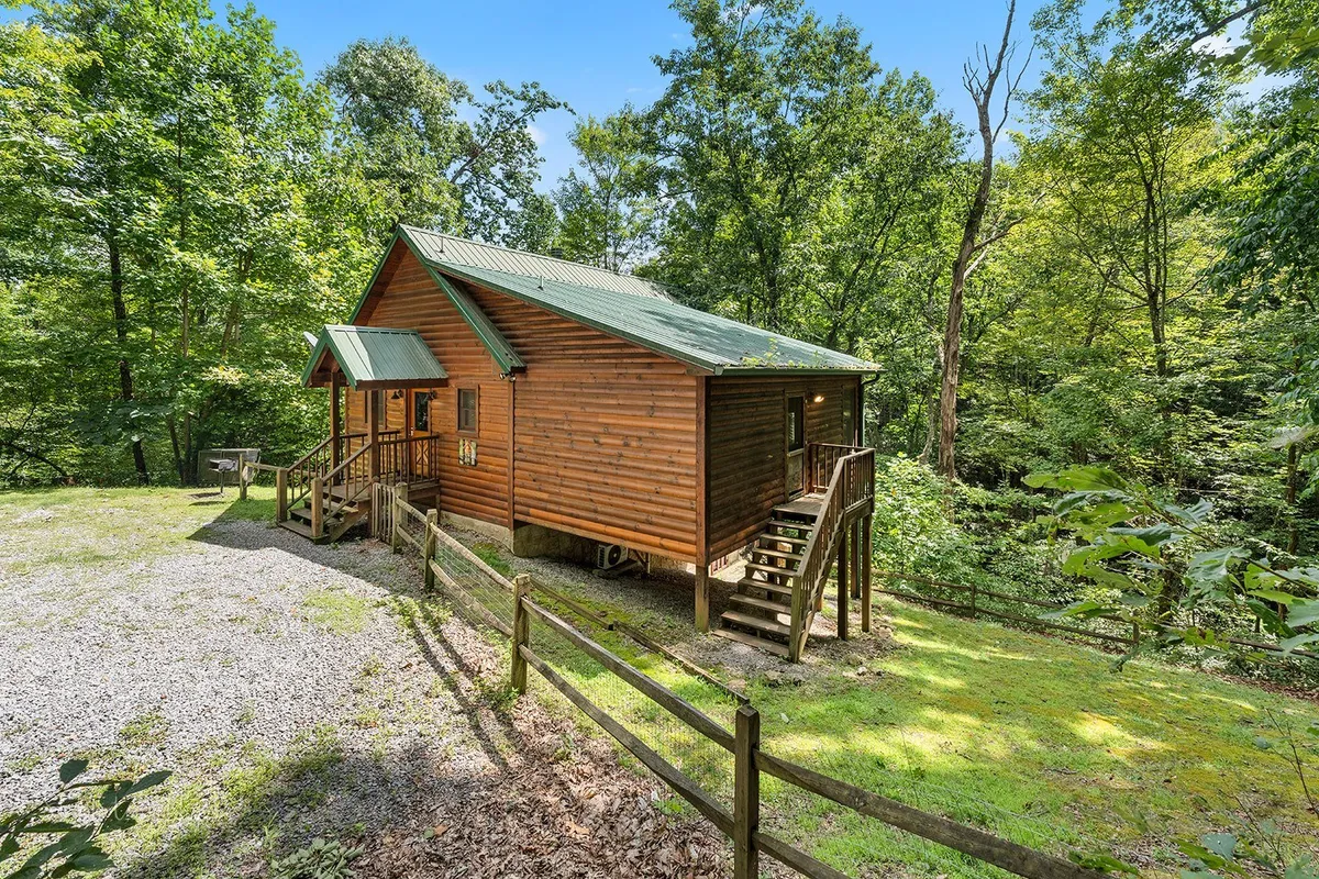 This exterior shot captures the wooden cabin amid the trees with a visible fenced area. The fencing adds a sense of security and practicality for guests bringing dogs, while the lush greenery and porch promote outdoor enjoyment and easy access to hiking trails.