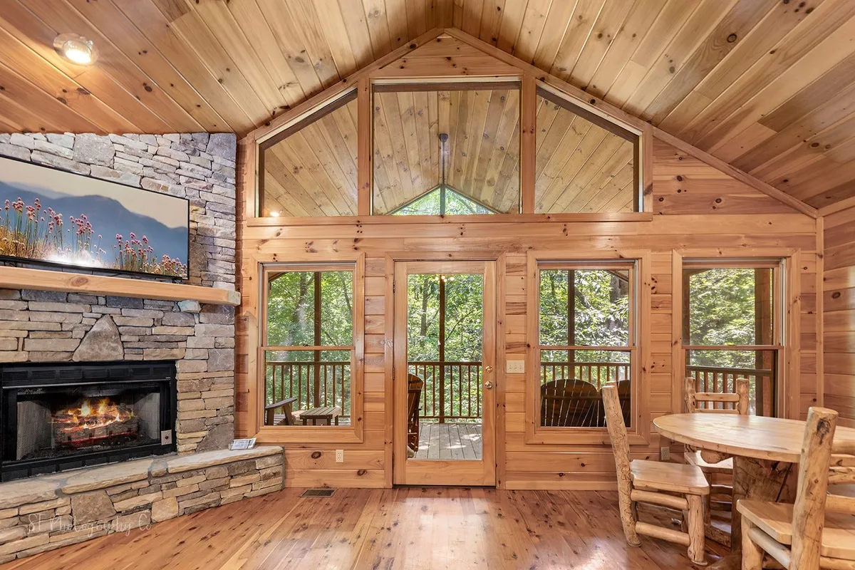 An open-plan space that combines a large wooden dining table, a fireplace, and expansive windows looking out to nature. The setting encourages family meals and gatherings, with the warmth of the fire enhancing the ambiance. Natural wood accents and ample sunlight create a bright, welcoming dining area.