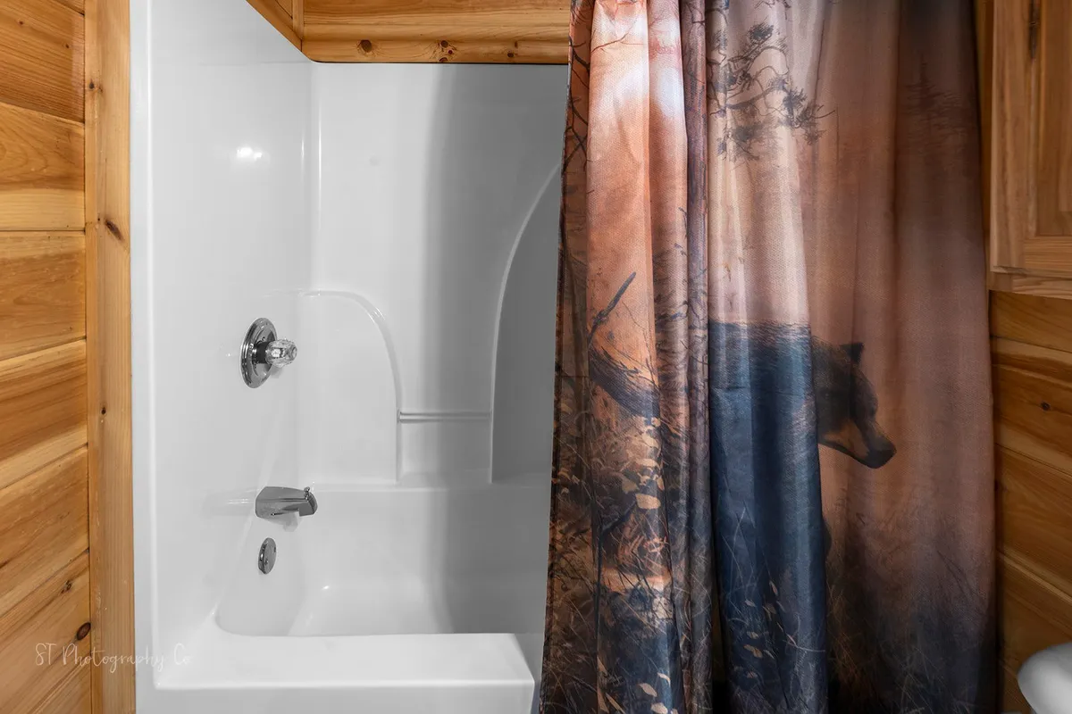 A tranquil bathroom featuring a bathtub with a bear motif shower curtain, wooden wall paneling, and soft lighting. The scene suggests a peaceful environment for unwinding after outdoor adventures.