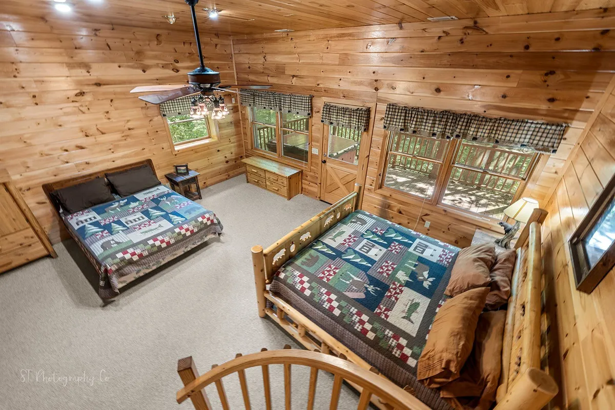 A tranquil bedroom setting with two large beds, wooden walls, a spiral staircase, and windows adorned with checkered curtains. The space exudes rustic charm and is designed for both relaxation and togetherness, with plenty of sleeping space and natural light.