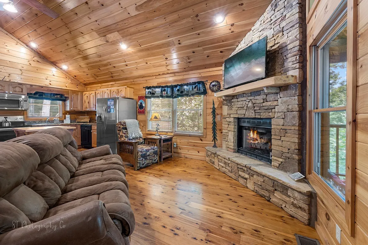 A cozy living room with a stone fireplace, plush couch, wooden ceiling, and a large window with mountain views. The space invites relaxation after a day of adventure and highlights the warmth and welcoming atmosphere of the cabin.