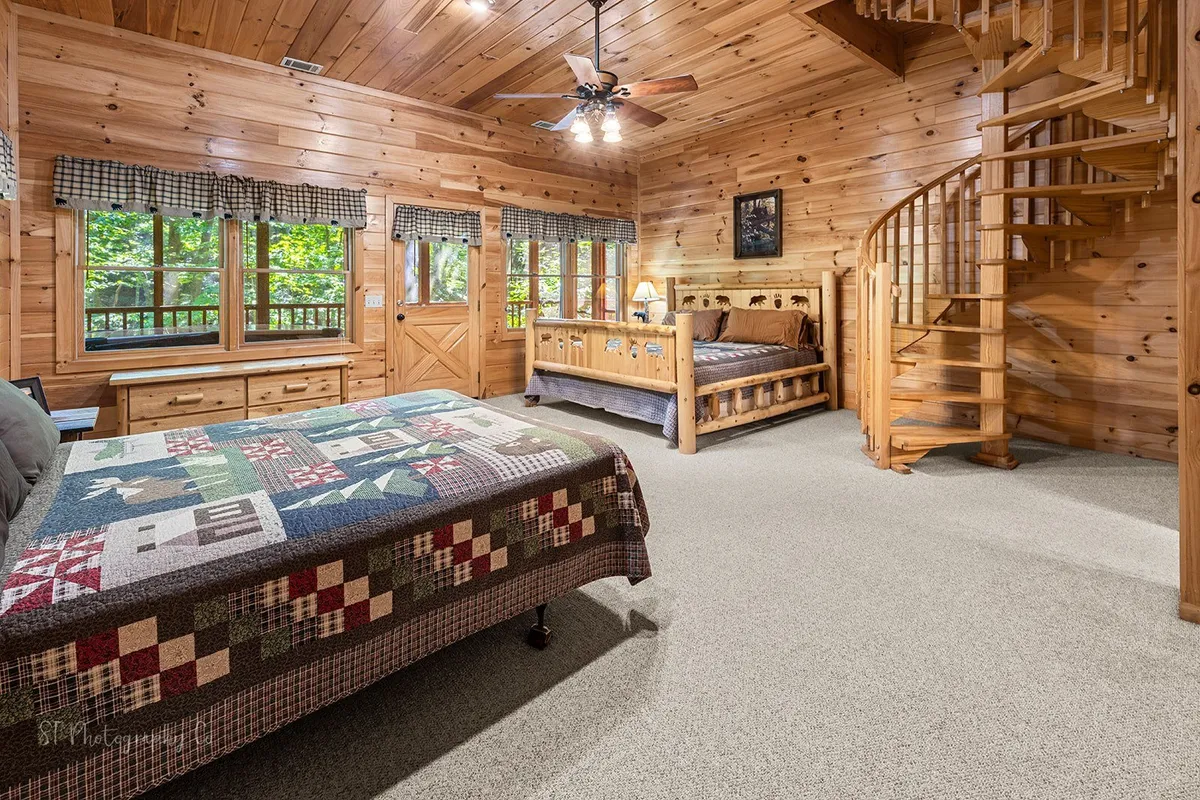 A bright room featuring a wooden spiral staircase, a quilted bedspread, and large windows that showcase lush outdoor scenery. The space is warm and welcoming, perfect for relaxing with family while appreciating the beauty of the seasons outside.