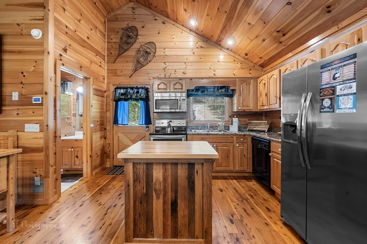 A well-appointed kitchen featuring stainless steel appliances, a kitchen island with additional seating, and rustic wooden cabinetry. The space is bright and functional, ideal for cooking, socializing over meals, or morning coffee. Blue curtains add a pop of color, while the overall look remains warm and homey.