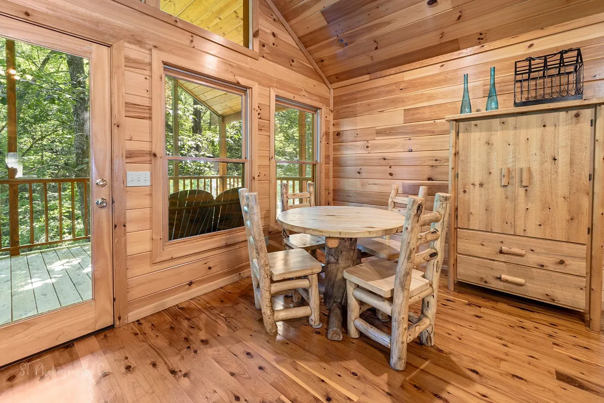 A rustic dining area with a wooden table and log chairs, surrounded by natural wood paneling and large glass doors framing an inviting view outside. The setting suggests lively meals, games, and conversations, all bathed in natural light.