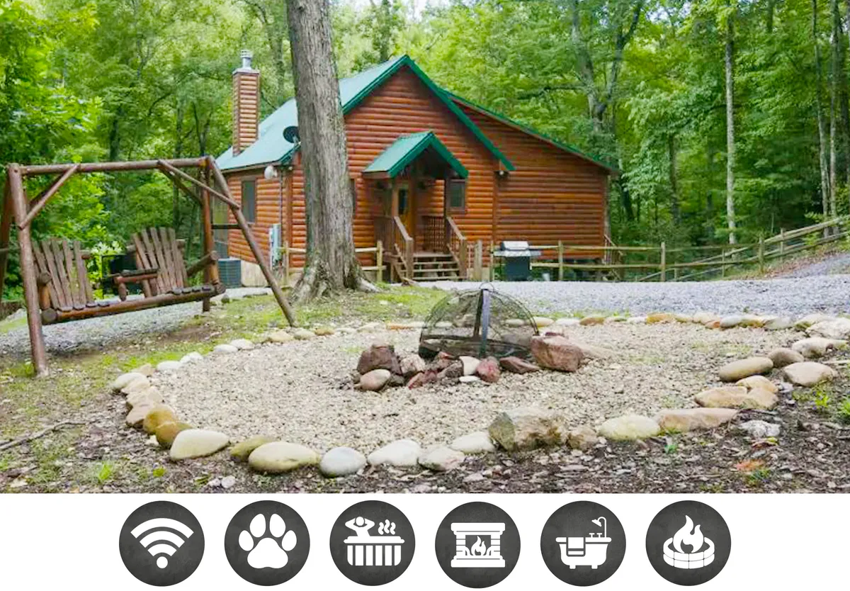 The image shows a charming log cabin nestled among lush trees in Wear's Valley, Tennessee. In the foreground, a cozy fire pit area is surrounded by seating, perfect for gathering under the stars. A rustic wooden swing hangs nearby, inviting guests to relax and soak in the tranquil mountain atmosphere.
