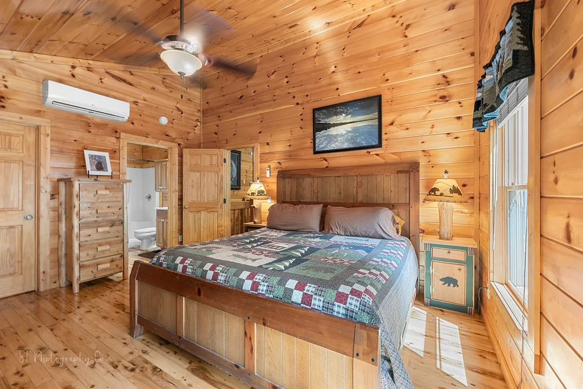 A cozy bedroom features a wooden bed with a patchwork quilt, a sturdy wooden dresser, and lamps with bear motifs. Natural light streams in, creating a warm and restful space ideal for relaxation after a busy day. The room includes closets and storage options, reflecting attention to comfort and practicality.