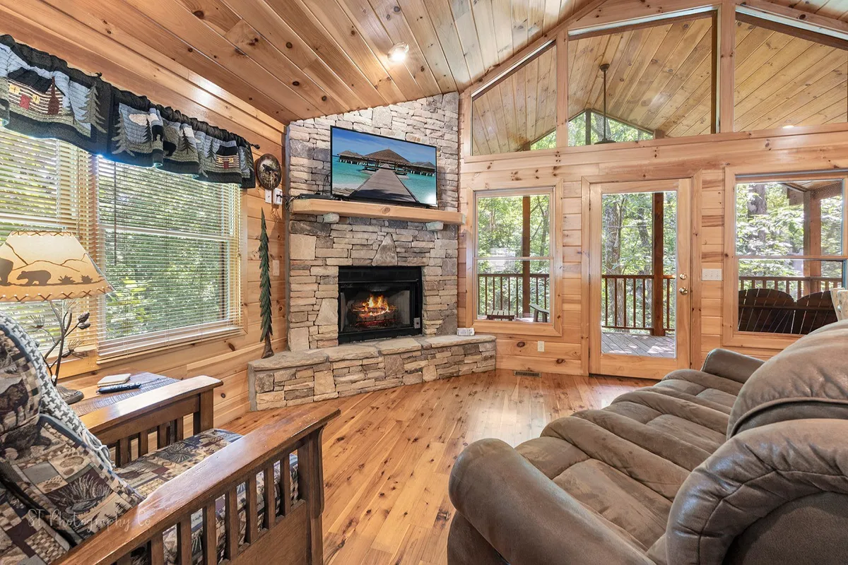 A cozy living room featuring a gas fireplace as the central focal point, flanked by large windows that fill the space with natural light. Plush, comfortable seating faces both the fireplace and a smart TV, inviting group gatherings or quiet relaxation. The warm wooden accents and mountain decor complete the inviting atmosphere.