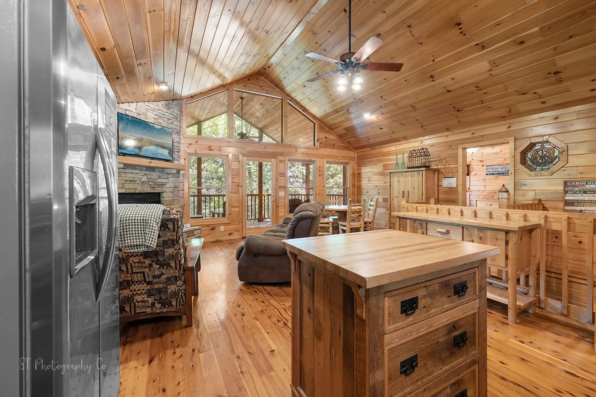 A spacious, inviting living area with a large stone fireplace, a comfortable armchair, and a wooden dining table. Sunlight pours in through expansive windows, offering views of the surrounding trees. The space is styled in warm wood tones and is ideal for gathering, relaxing by the fire, or enjoying the view.
