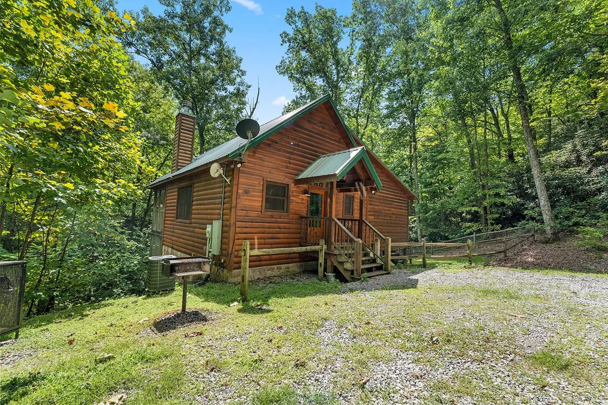 This image shows the log cabin's exterior nestled among mature trees, with a green roof and gravel driveway. The natural setting highlights the property's secluded, wooded environment, perfect for relaxing outdoors with pets. The front porch features comfortable seating, inviting guests to enjoy time outside and take in sunset views through the trees.