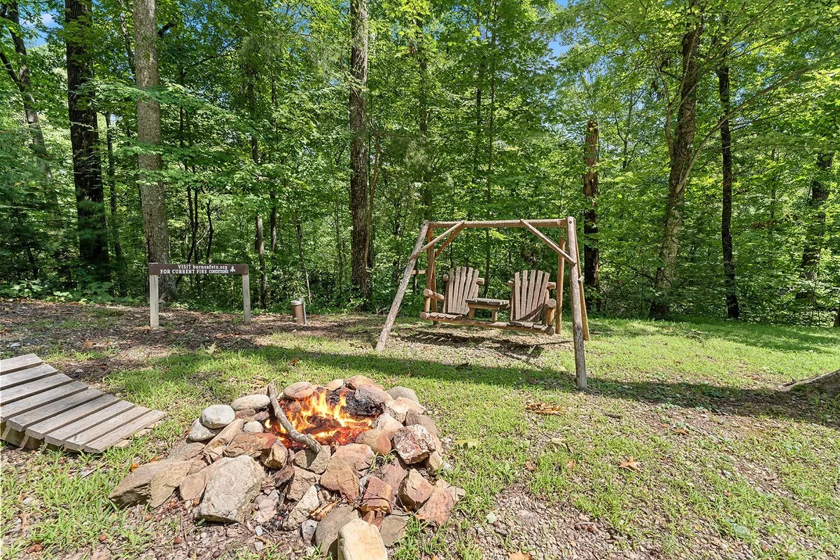 A tranquil exterior view of the fire pit area surrounded by trees, with a swing set and rustic seating. The scene evokes the serenity of evenings spent outdoors, enjoying nature and each other's company under the open sky.
