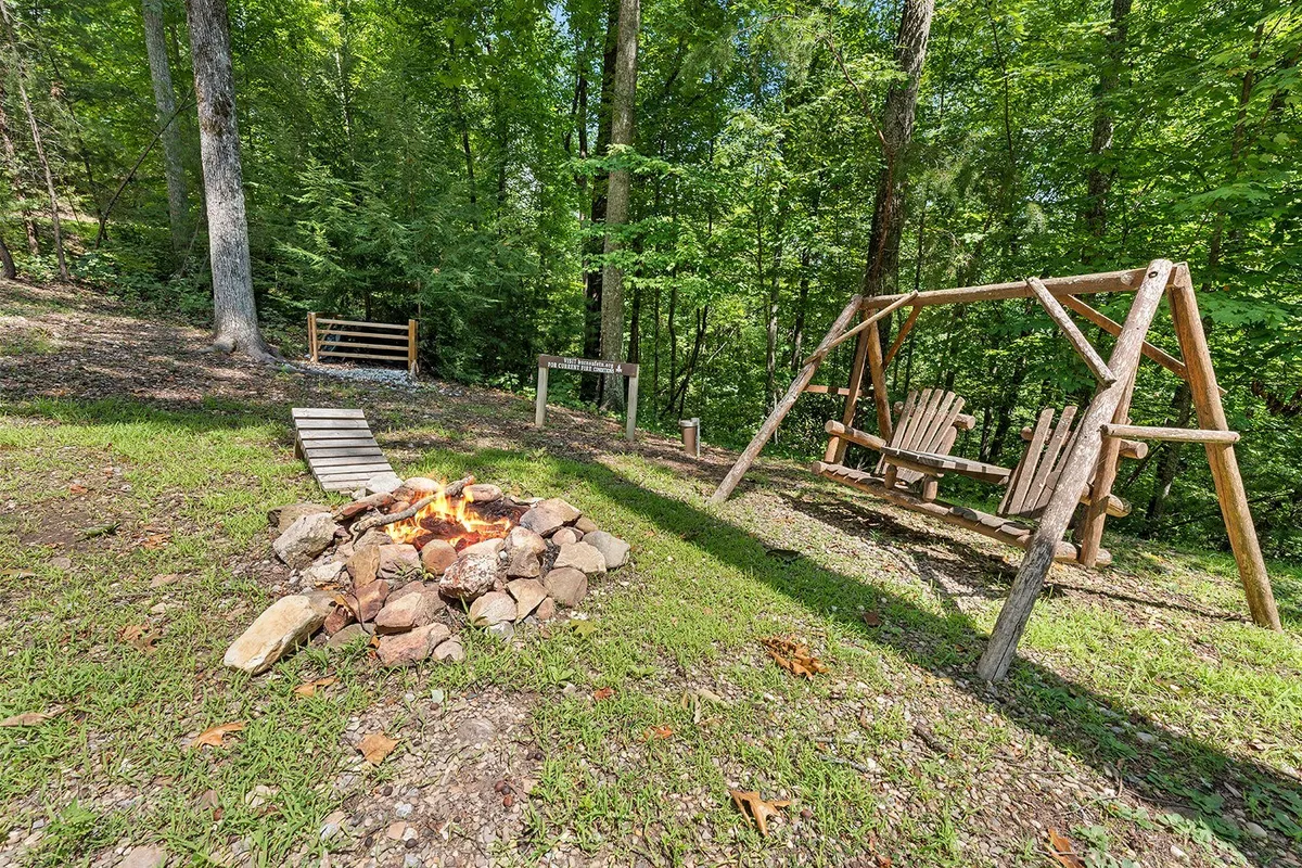 An inviting outdoor space featuring a fire pit surrounded by chairs and a swing set, nestled within lush greenery. The scene exudes warmth and community, with the fire pit ready for s’mores and stories while the swing set offers another way to enjoy the peaceful environment.